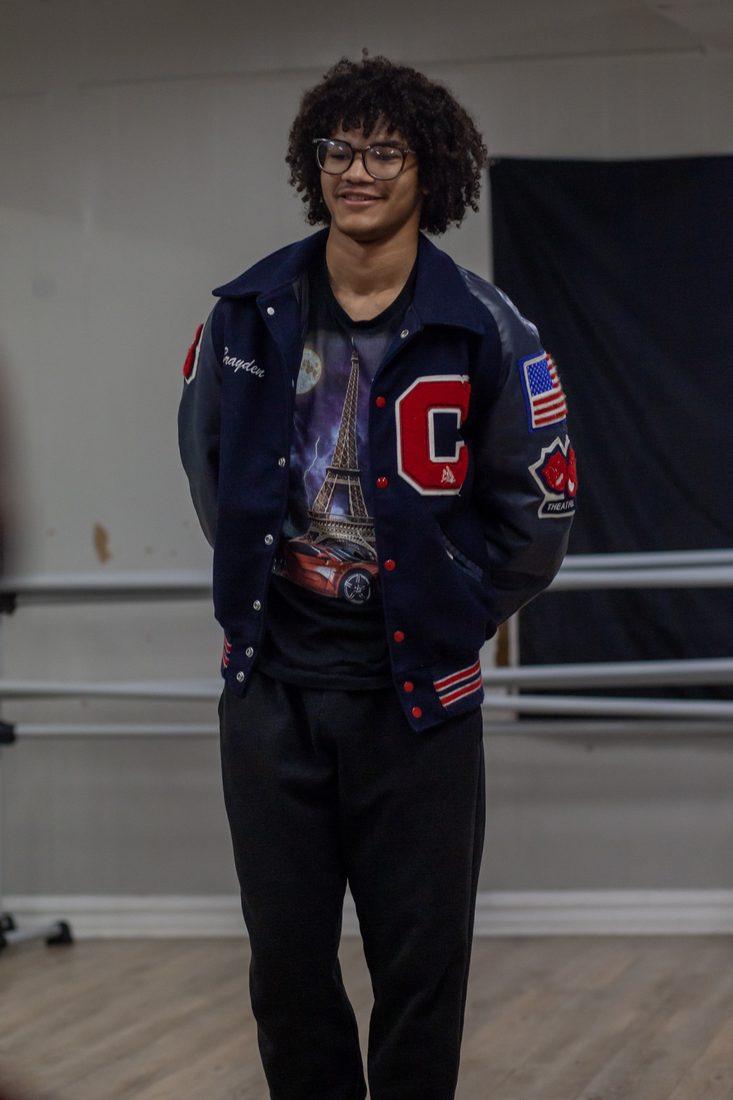 Brayden Landry standing in a dance studio, smiling, in a navy letterman jacket with a red C